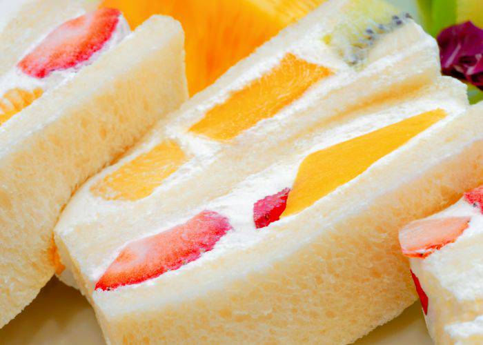 A close-up of fruit sandwiches: colorful strawberries and mangos, fluffy cream and white bread.
